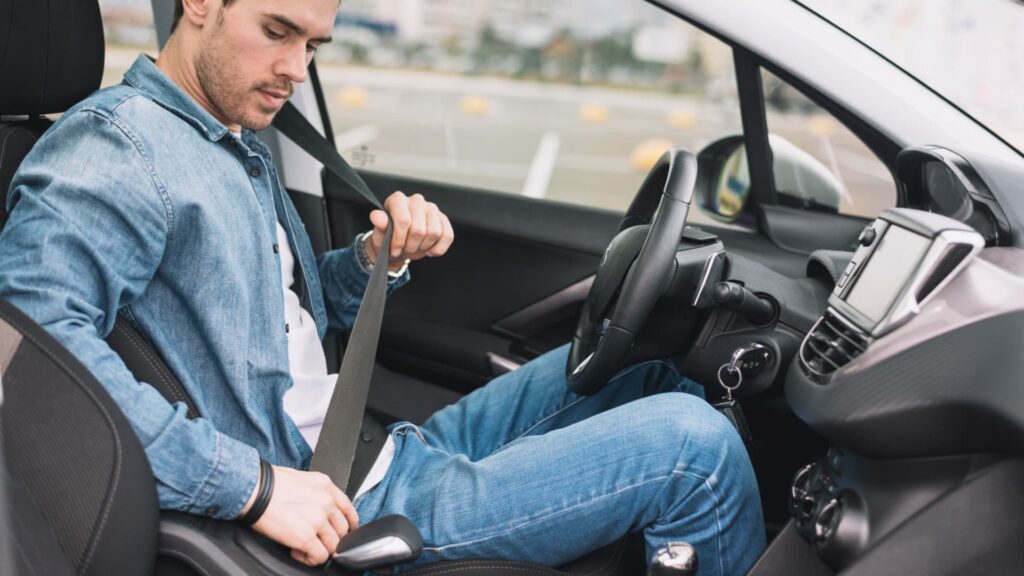 Homem com jaqueta jeans botando um cinto de segurança preto em um carro com a interna preta.