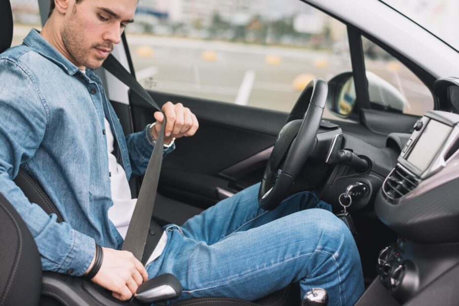 Homem com jaqueta jeans botando um cinto de segurança preto em um carro com a interna preta.