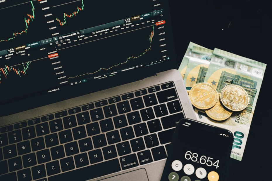 Notebook aberto em uma plataforma de criptomoedas com um celular aberto na calculadora e moedas de ouro em cima de notas de dinheiro.