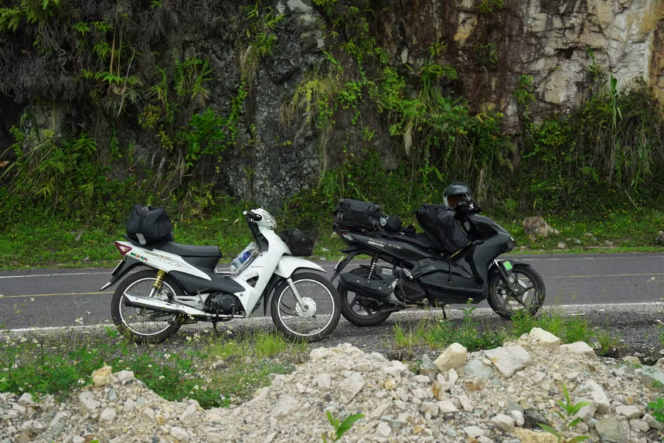 Duas motocicletas estacionadas na beira da estrada com um penhasco rochoso e vegetação verde ao fundo